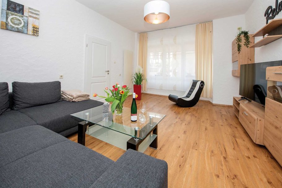 Moderne Wohnzimmer mit schwarzem Sofa, Glastisch, Fernseher und hellen Vorhängen.